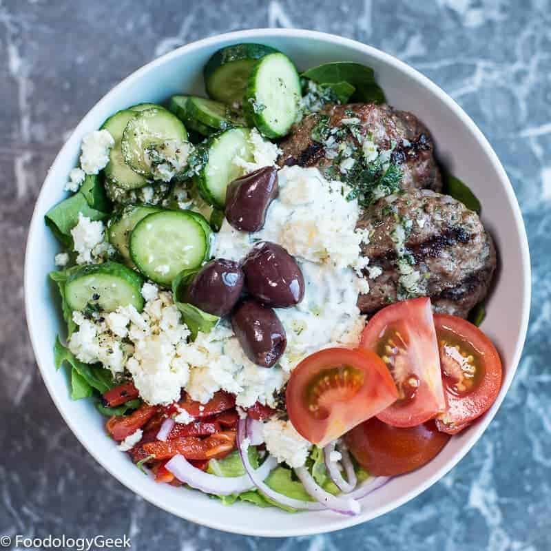 Mediterranean meal prep bowl. Greek mini burgers served on top of fresh vegetables. Served with greek yogurt dip, kalamata olives, feta, and Greek dressing.