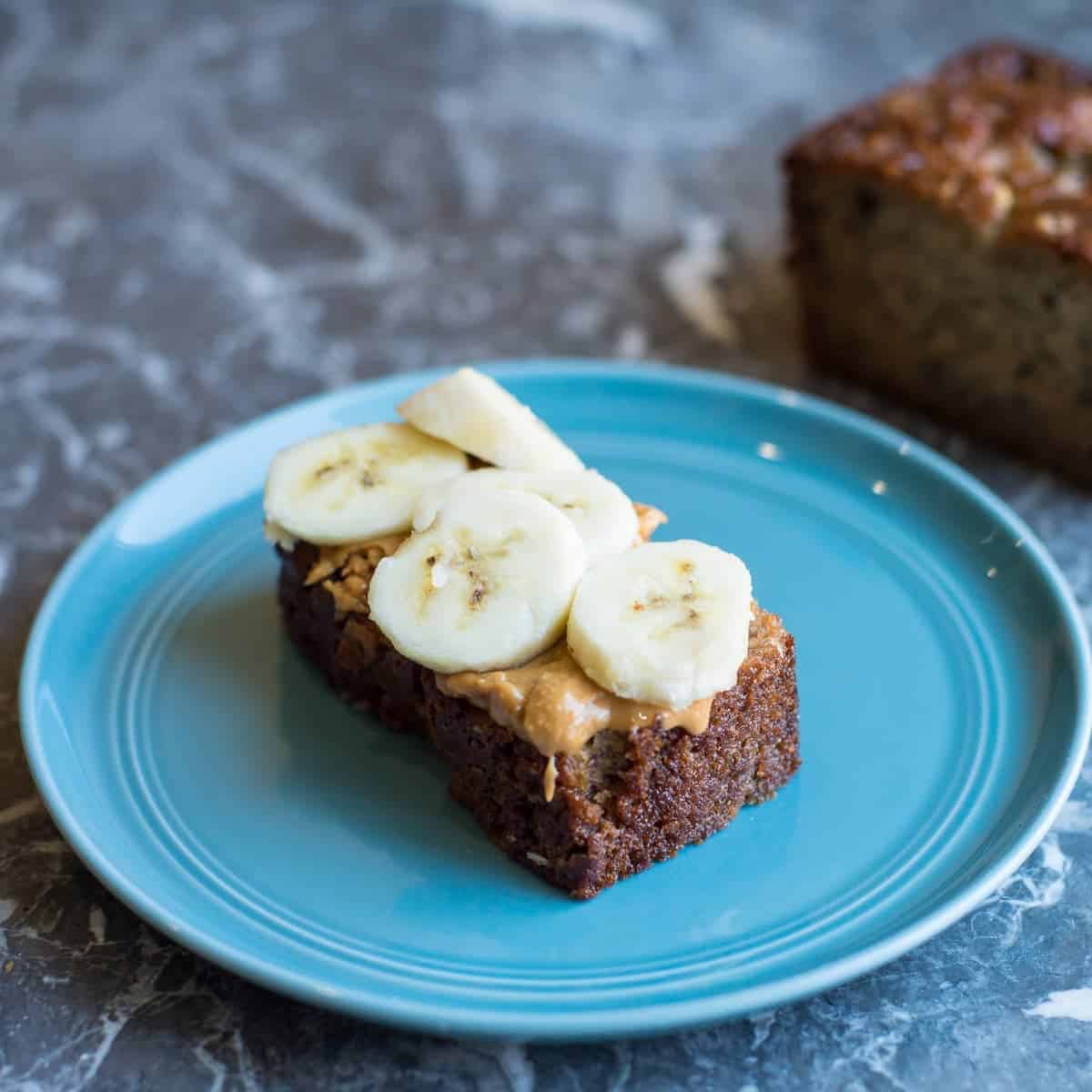 A slice of banana bread slathered with peanut butter and topped with freshly sliced bananas by foodology geek