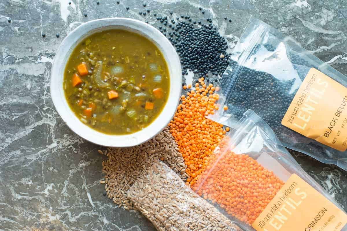 Lentil vegetable soup recipe in a bowl. Surrounded by black belug lentils, red lentils, and farro by foodology geek.