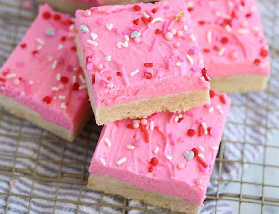 A soft sugar cookie bar decorated with pink frosting and sprinkles. #SugarCookies #BarreCipes Stack of sugar cookie bars decorated with pink icing and Valentine's Day sprinkles.