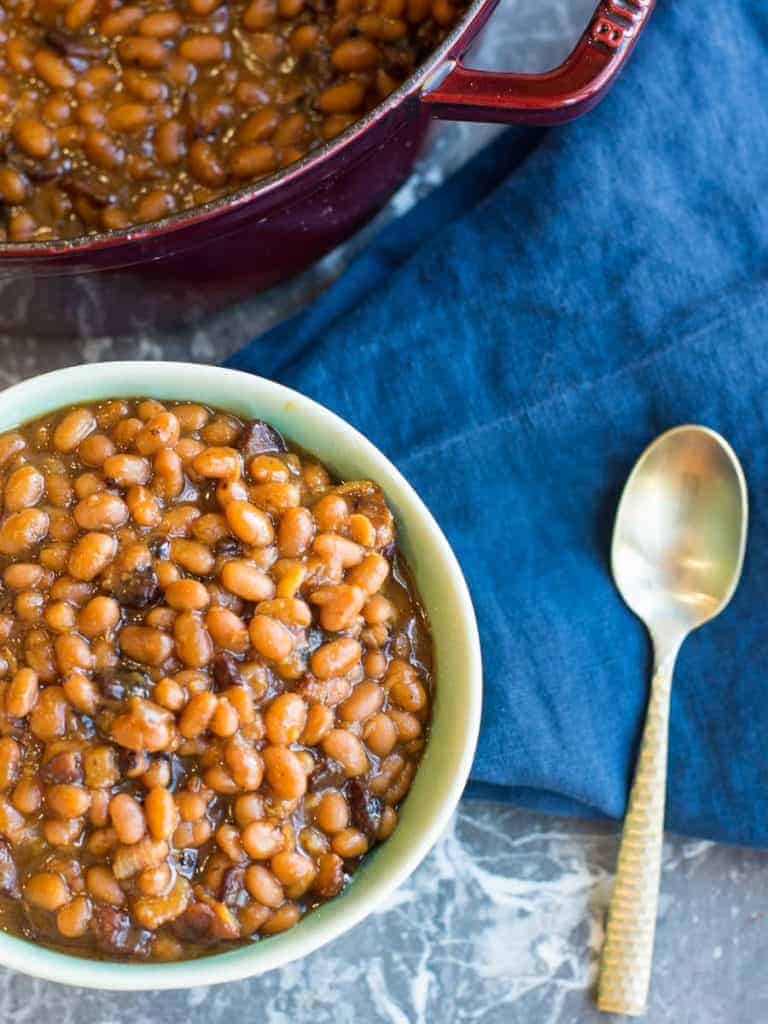 a bowl of baked beans with brown sugar and bacon, gold spoon and a staub cast iron pan