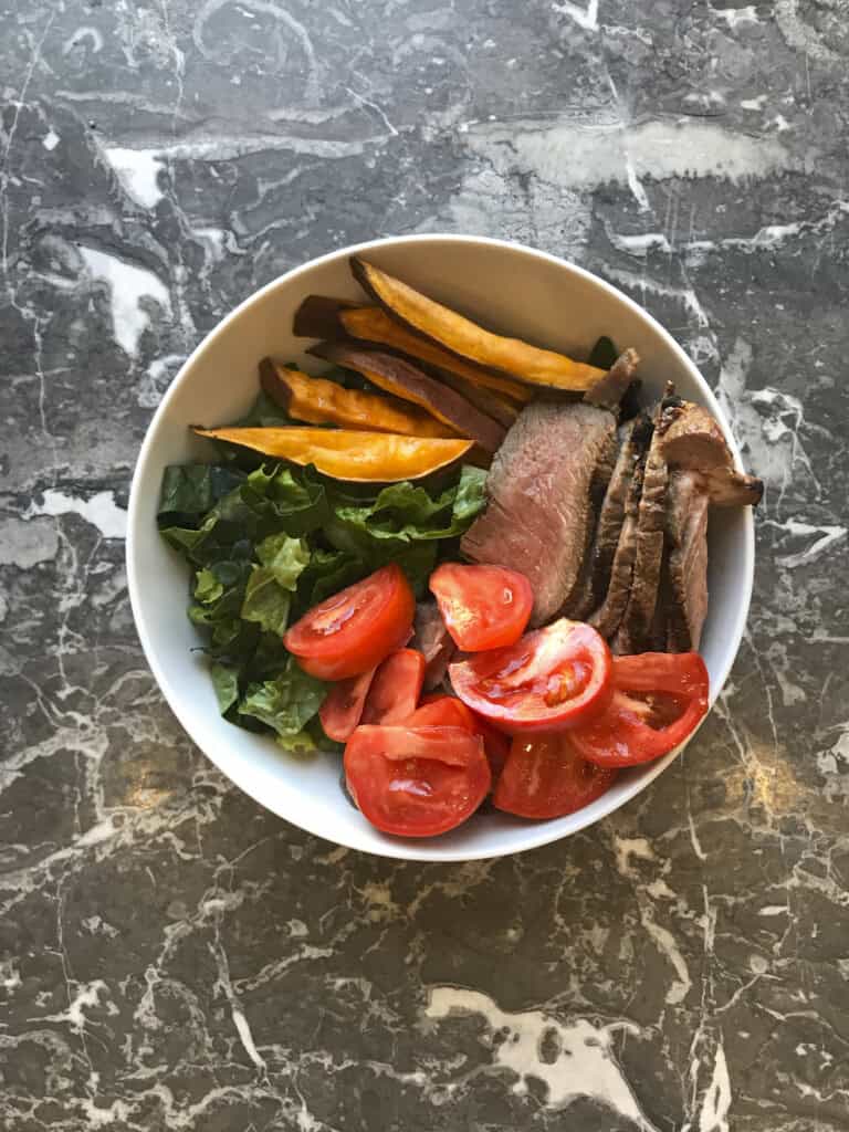 steak and sweet potato meal prep bowl with greens, steak and sweet potato fries and tomatoes