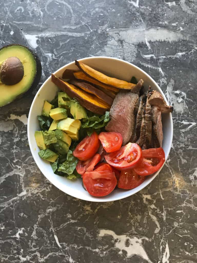steak and sweet potato meal prep bowl with greens, steak and sweet potato fries and tomatoes, plus avocado
