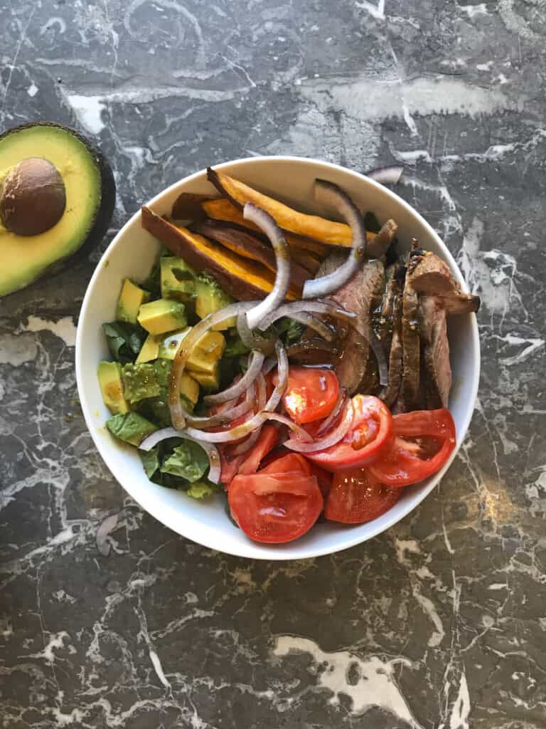 steak and sweet potato meal prep bowl with greens, steak and sweet potato fries and tomatoes, plus avocado and red onions