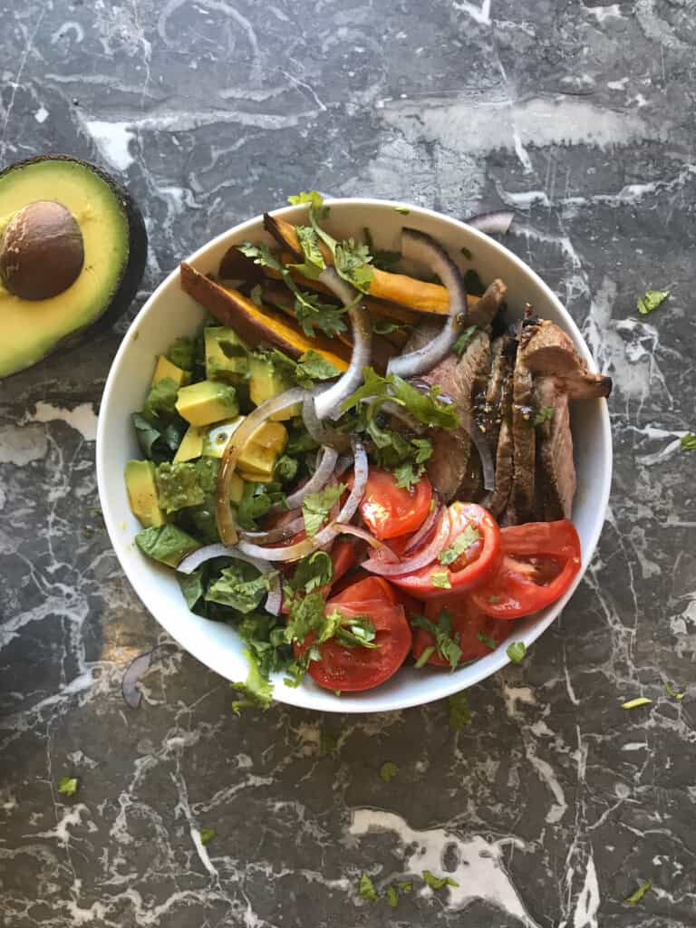 steak and sweet potato meal prep bowl with greens, steak and sweet potato fries and tomatoes, plus avocado and red onions, drizzled with balsamic vinaigrette